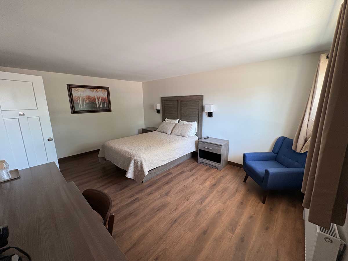 Renovated queen room with modern flooring, queen bed, comfortable armchair, work desk, mini fridge, and microwave.